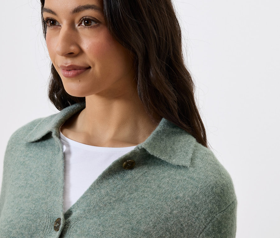 Porträt einer Frau mit dunklem Haar, die einen grünen Pullover mit Kragen und Knöpfen trägt.