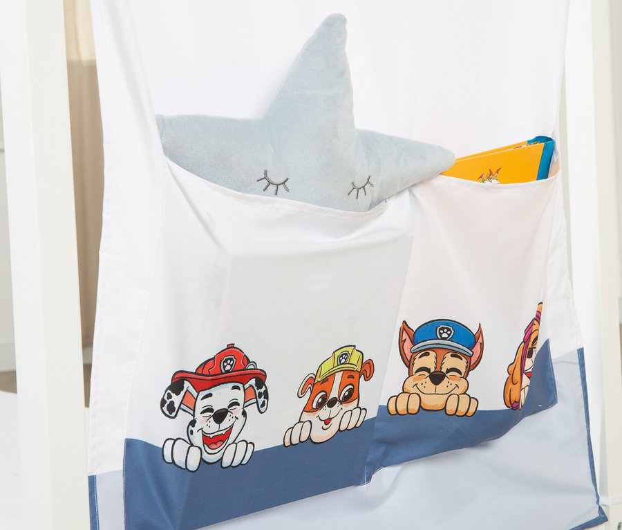 Eine weiße Stofftasche mit Paw Patrol-Figuren hängt an einem Bett. In den Taschen befinden sich ein blaues Sternkissen und Bücher.