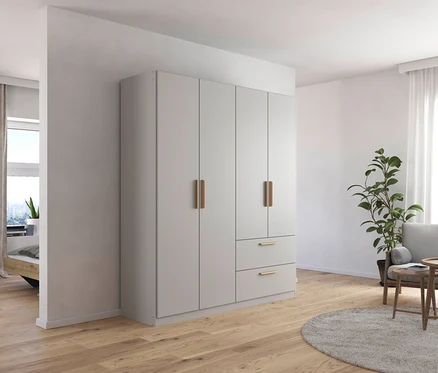 Ein Zimmer mit einem 4-türigen, seidengrauen Drehtürenschrank »Skandi« von Rauch, einem Sessel, einer Pflanze und einem Holzpodest.