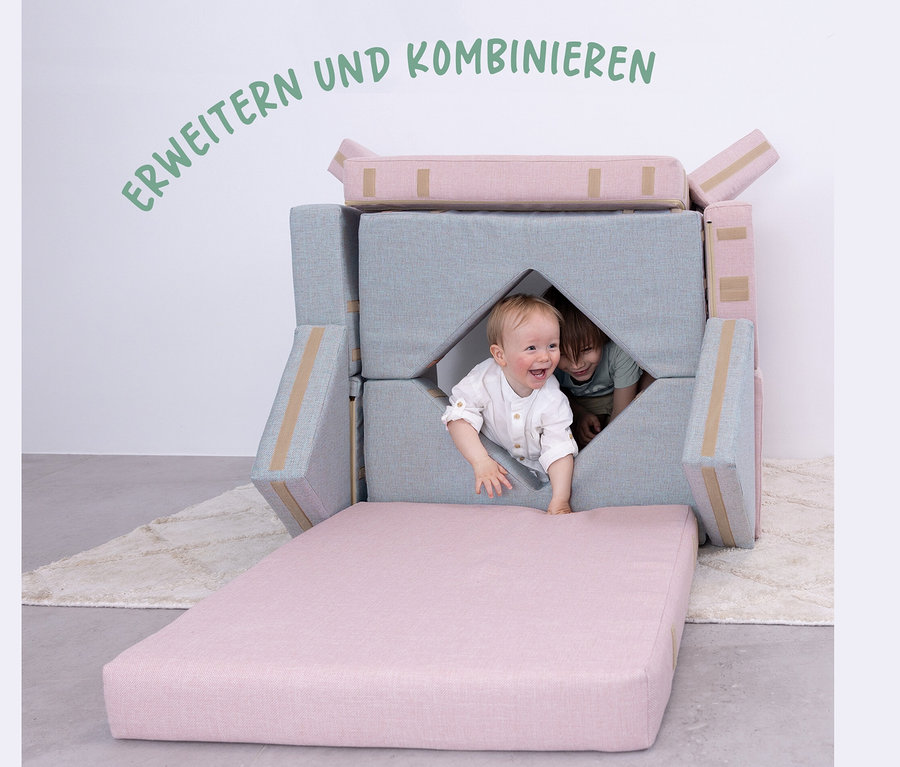Ein Kind klettert aus einem Buddies Spielsofa, mintgrau und rosa, während ein anderes Kind aus einem Loch späht.