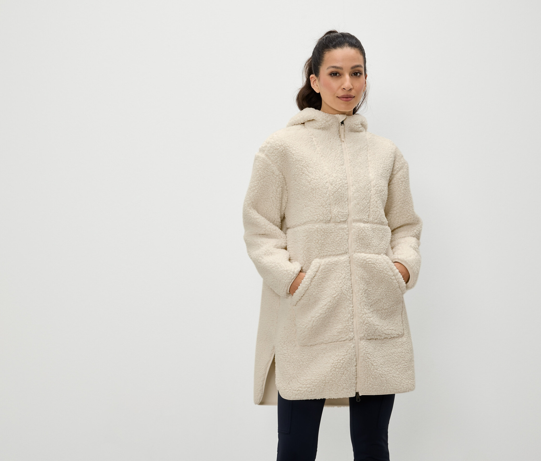 Frau steht mit Händen in den Taschen eines cremefarbenen, gefütterten Mantels mit Reißverschluss und Kapuze.