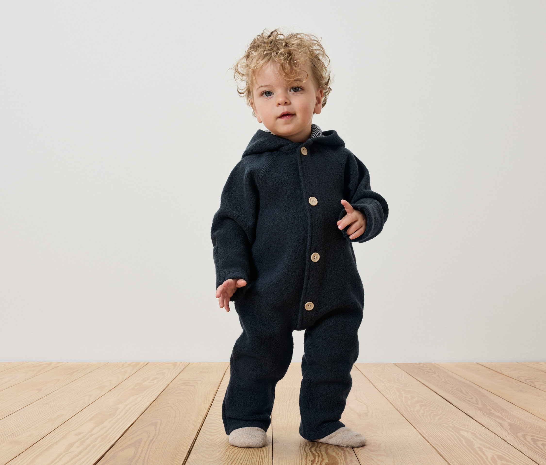 Ein Kind mit lockigem Haar steht auf einem Holzboden und trägt einen dunkelblauen Overall mit Kapuze und Knöpfen.