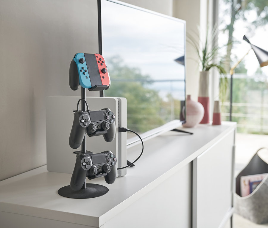 Auf einer weißen Kommode steht ein schwarzer YAMAZAKI Controller-Ständer »SMART« mit einem Nintendo Switch-Controller und zwei schwarzen Gamecontrollern.