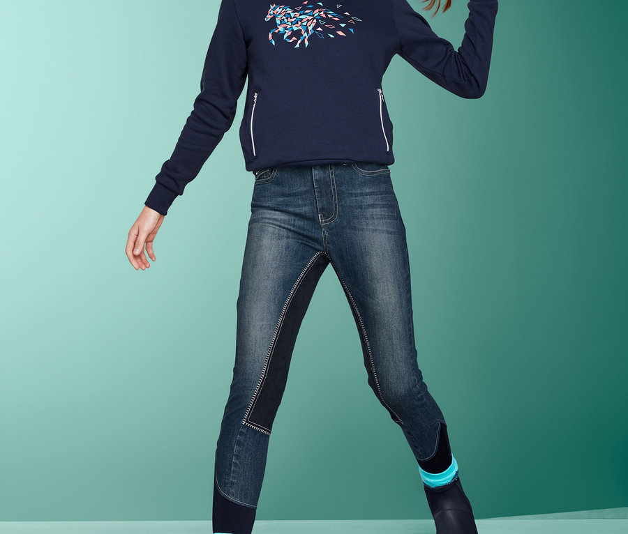 Ein junges Mädchen in einem dunkelblauen Hoodie und einer USG-Jeans-Reithose posiert vor einem grünen Hintergrund.