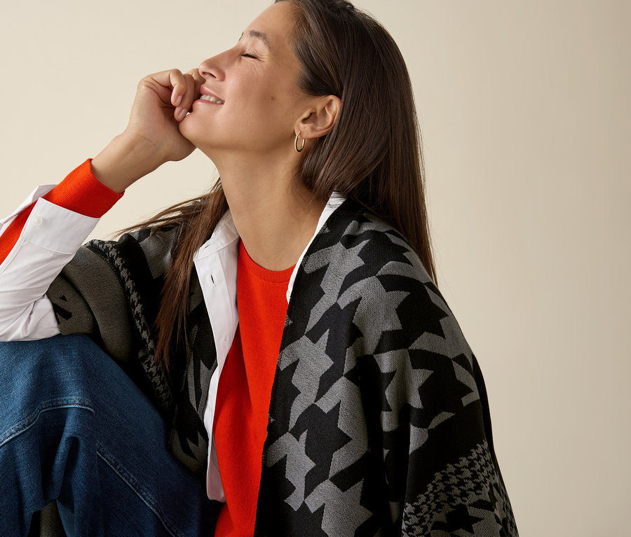 Frau sitzt und trägt einen orangen Mernio-Feinstrickpullover.