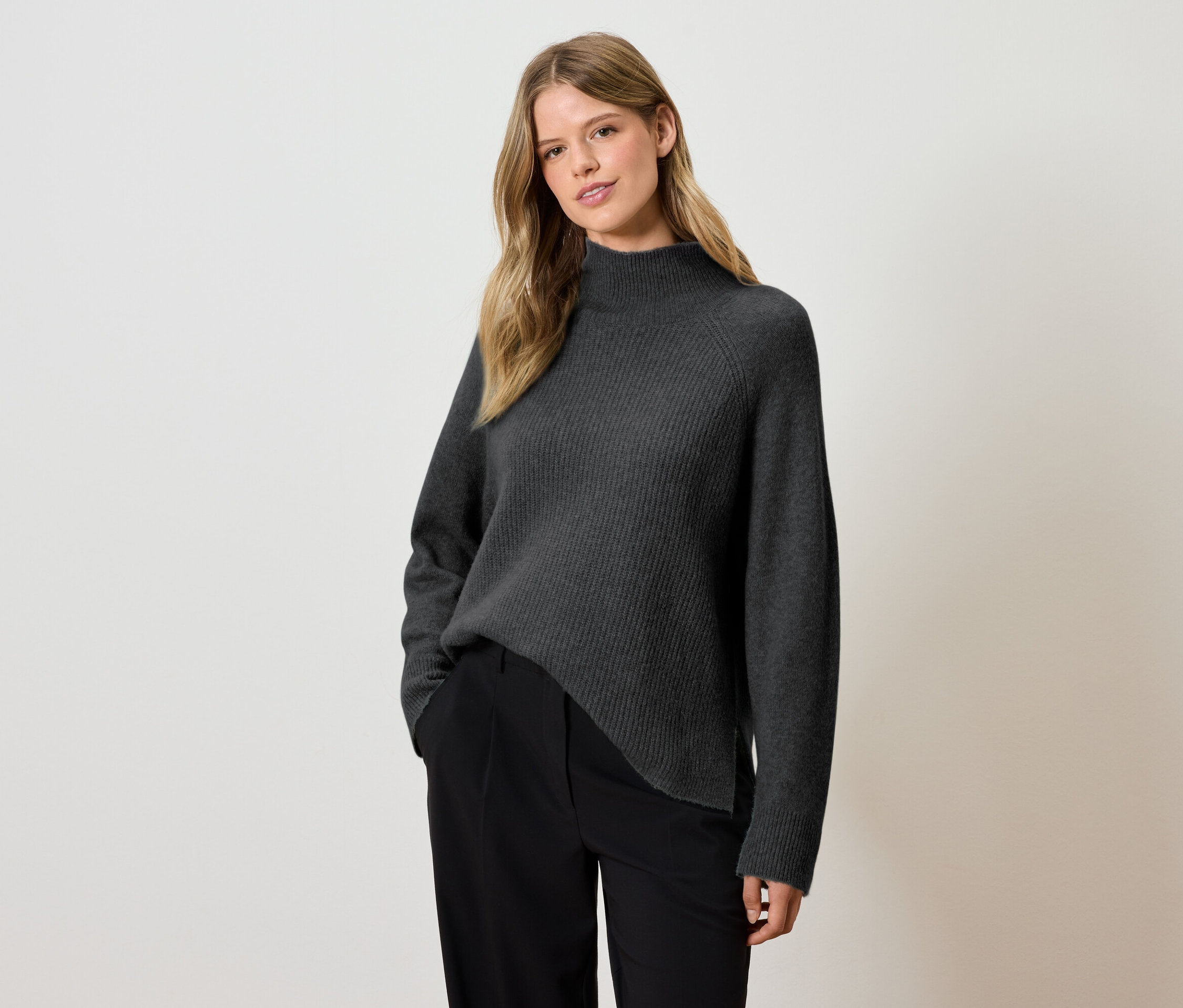 Frau posiert in einem grauen Pullover und einer schwarzen Hose.