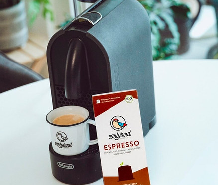 Eine Tasse Kaffee steht auf einer DeLonghi Maschine, daneben eine Packung earlybird Bio Espresso Kapseln.