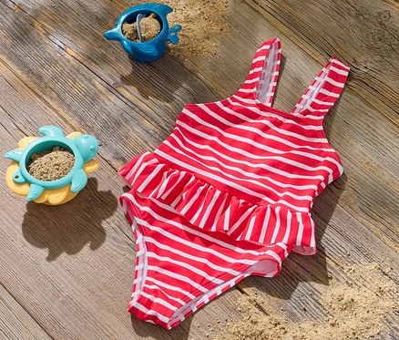 Ein Kinder-Badeanzug mit roten und weißen Streifen liegt auf einer Holzoberfläche neben zwei Sandspielzeugen.