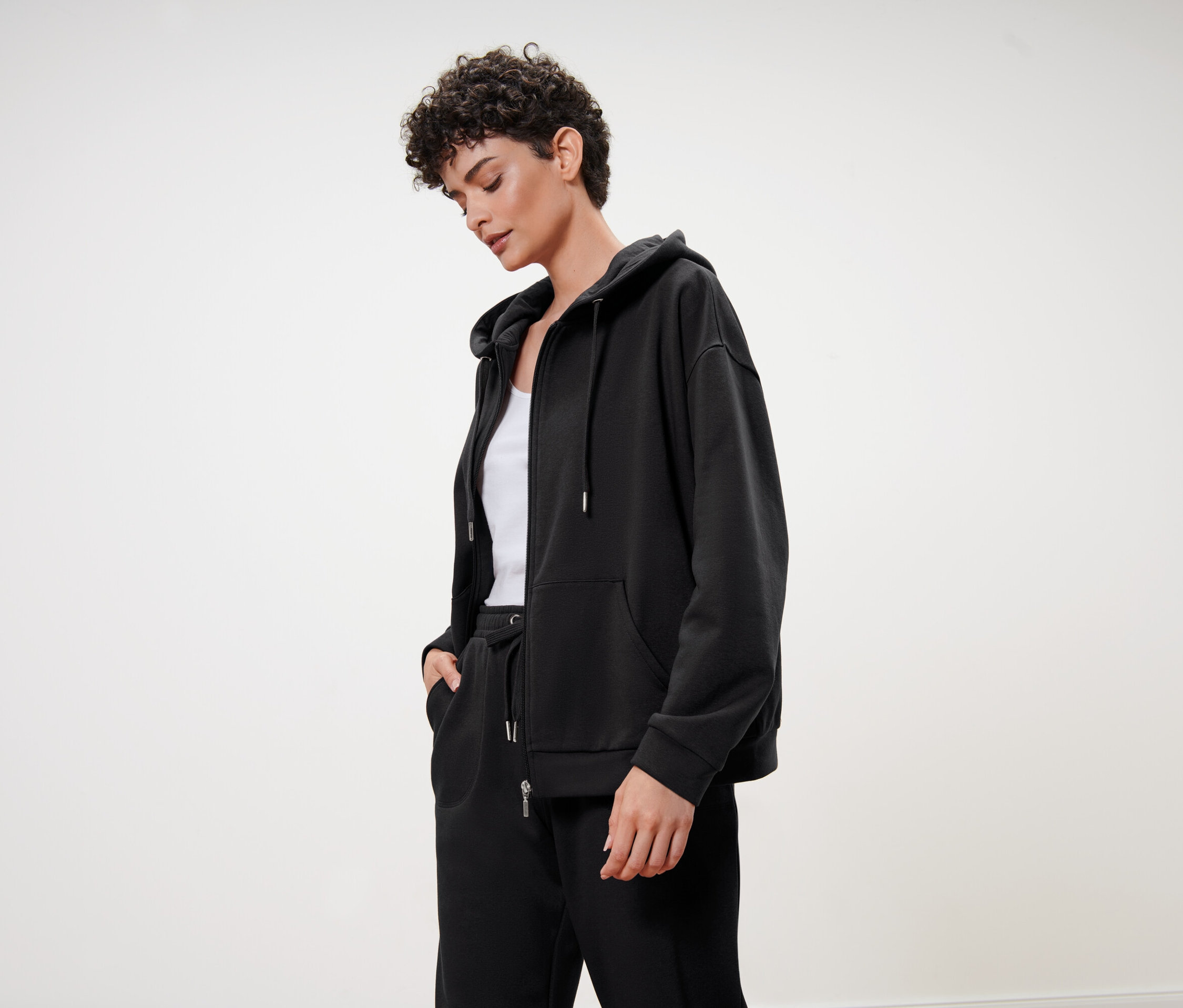 Frau mit kurzen lockigen Haaren trägt einen schwarzen Kapuzenpullover, eine schwarze Trainingshose und ein weißes Top.