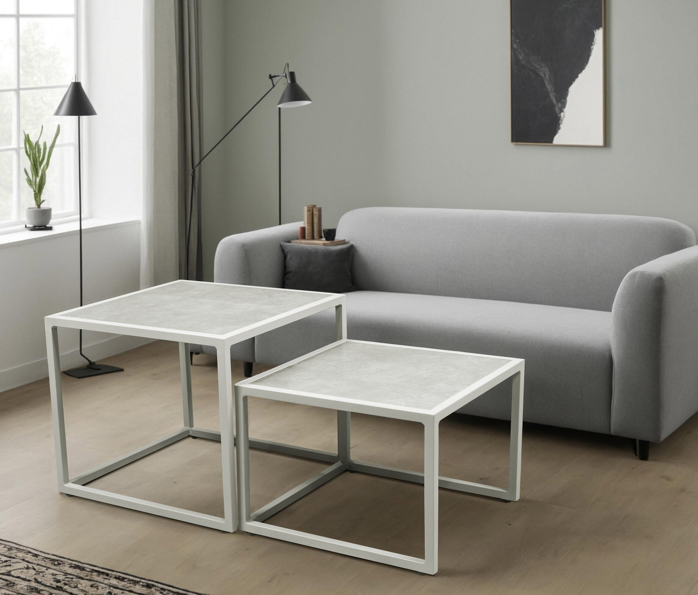 Zwei quadratische, weiß umrandete Couchtische mit grauer Platte stehen vor einem grauen Sofa in einem Wohnzimmer.