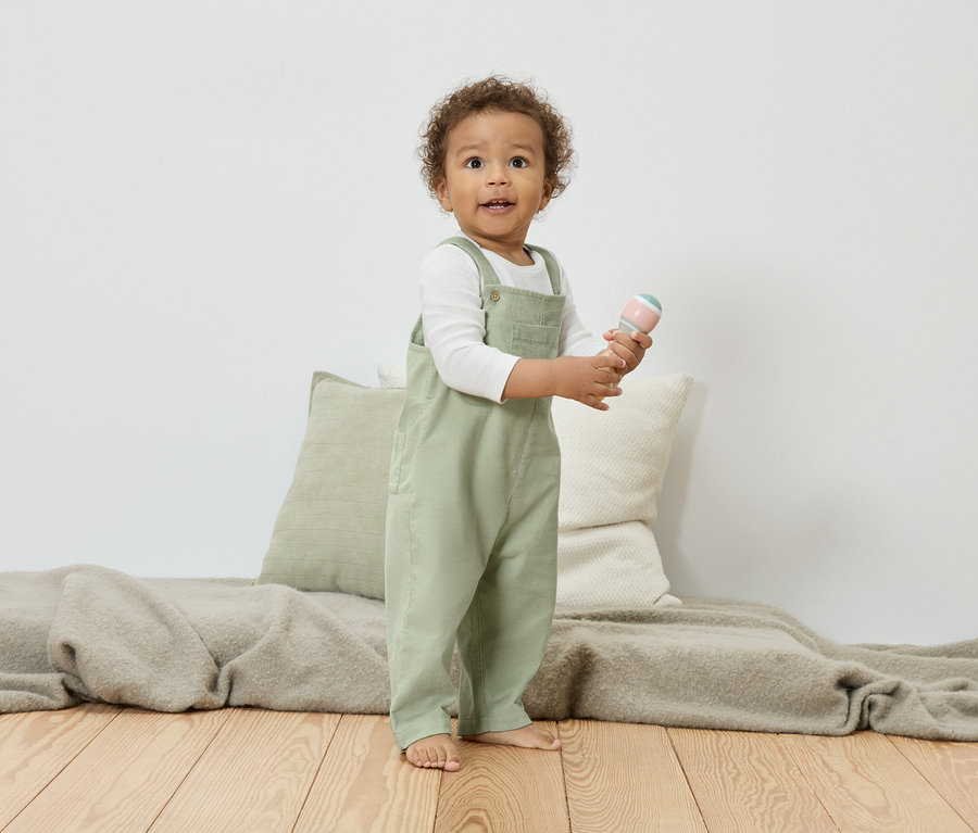 Ein Kleinkind mit lockigem Haar steht auf einem Holzboden und hält eine Rassel. Das Kind trägt eine grüne Latzhose über einem weißen Langarmshirt.