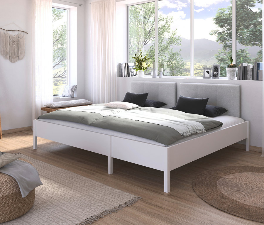 Weißes Doppelbett mit grauer Bettwäsche vor Fenster. Bücher und Dekoration auf Fensterbrett.