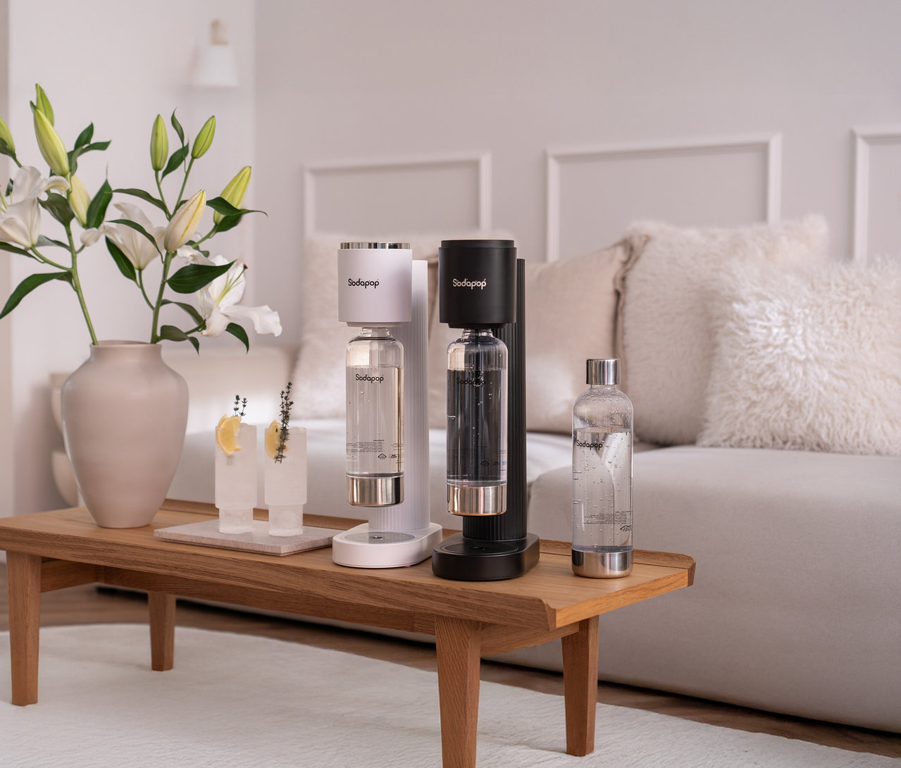Auf einem Tisch stehen zwei Sodapop Wassersprudler Cooper Pro, inkl. 5 Flaschen, in Weiß und Schwarz, neben Gläsern und einer Vase mit Blumen.