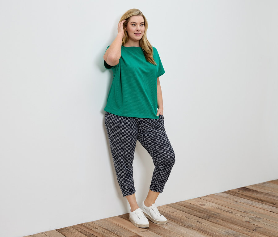 Frau mit grüner Bluse und gemusterter Hose lehnt an einer weißen Wand.