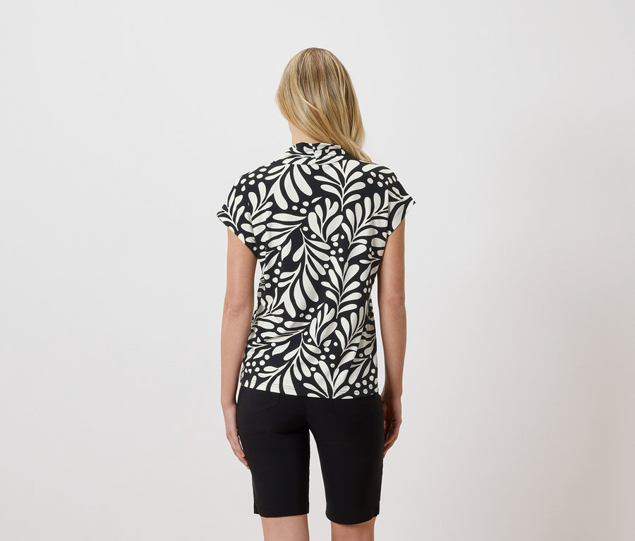 Rückenansicht einer Frau mit blonden Haaren, die eine schwarz-weiße Bluse mit Blätterdruck und schwarze Shorts trägt.