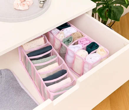 Ein offener Schubladenschrank zeigt zwei rosa Stoff-Organizer mit mehreren Fächern, gefüllt mit zusammengerollter Kleidung, und ein graues T-Shirt ist teilweise sichtbar.