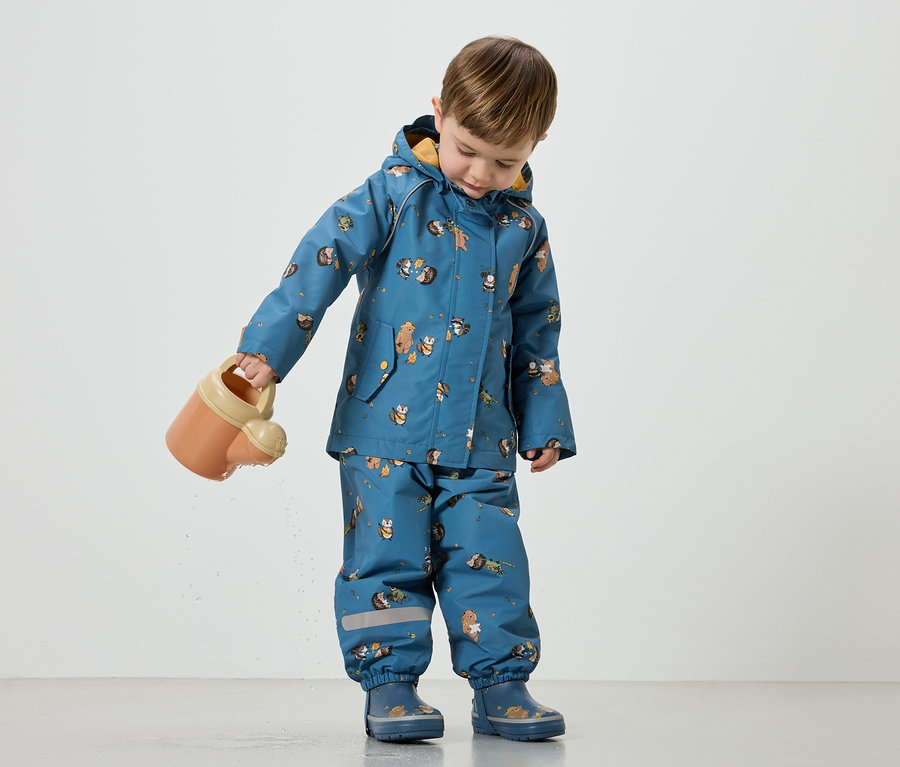 Ein Kind gießt mit einer orangefarbenen Gießkanne Wasser. Es trägt einen blauen Regenanzug mit Tiermotiven und passende Gummistiefel.