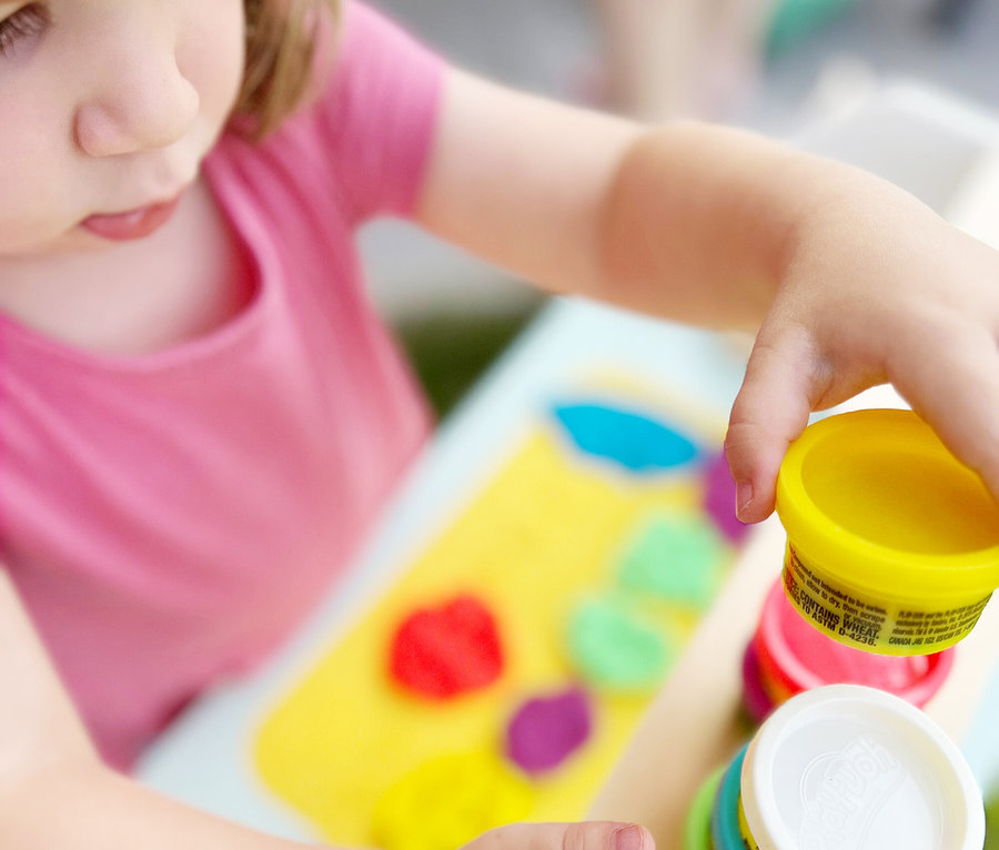 Ein Kind im rosafarbenen Hemd spielt mit gelbem, rotem, grünem und blauem Spielteig auf einem Tisch.