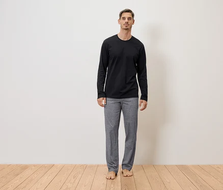 Mann steht barfuß in grauer Hose und schwarzem Langarmshirt vor einer Wand auf einem Holzboden.