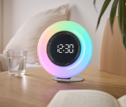 Auf einem Nachttisch steht eine bunte Lichtwecker mit der Uhrzeit 12:30, neben einem Glas Wasser und einem Buch.