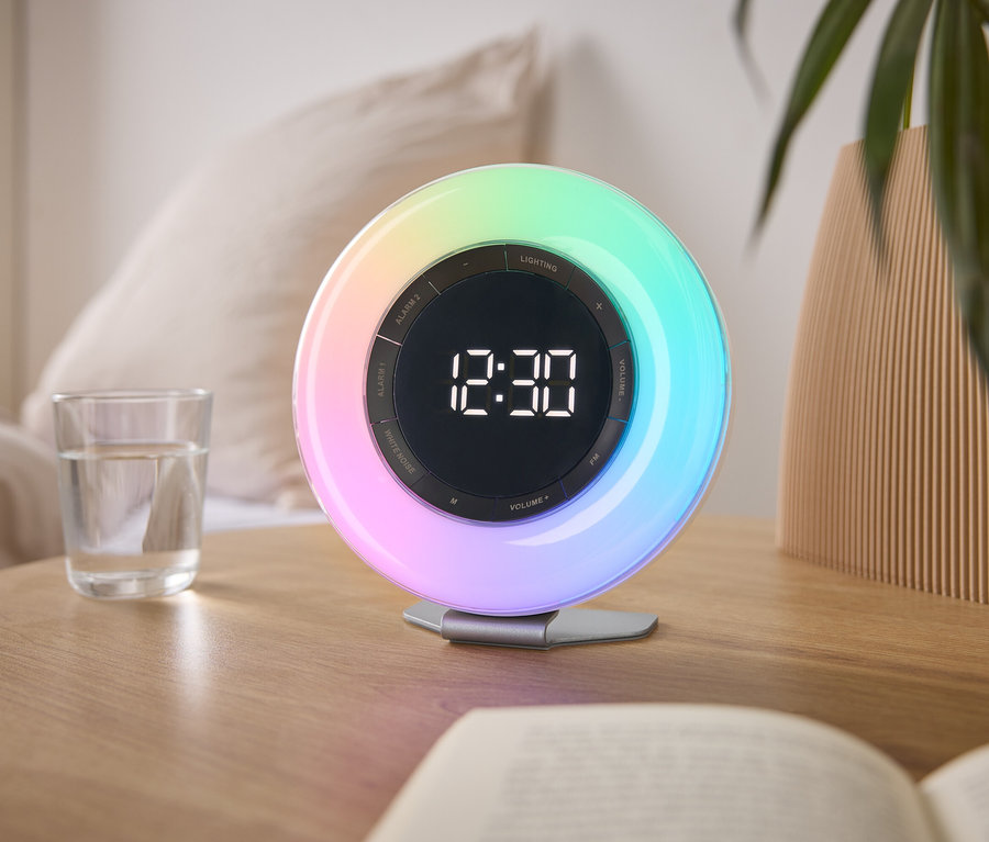 Auf einem Nachttisch steht eine bunte Lichtwecker mit der Uhrzeit 12:30, neben einem Glas Wasser und einem Buch.
