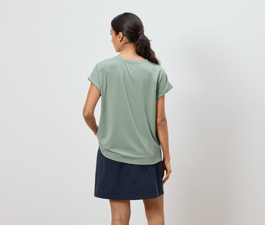 Frau von hinten in olivgrünem T-Shirt und blauem Rock vor weißem Hintergrund.