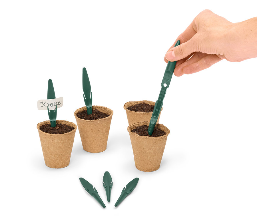 Eine Hand setzt einen grünen Samenstab in einen braunen Topf mit Erde, umgeben von weiteren Töpfen und Samenstäben.