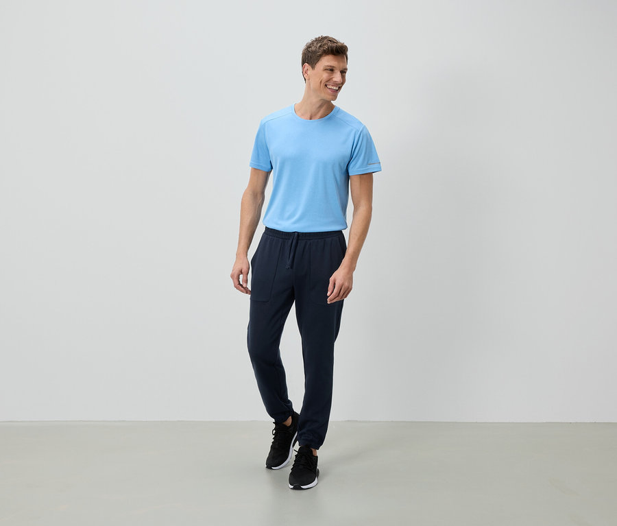 Ein lächelnder Mann steht in einem hellblauen T-Shirt, marineblauen Jogginghosen und schwarzen Turnschuhen vor einer weißen Wand.