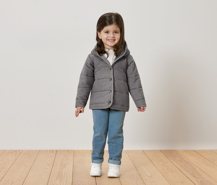 Ein kleines Mädchen mit braunen Haaren lächelt in die Kamera und trägt eine graue Jacke, Jeans und weiße Turnschuhe.