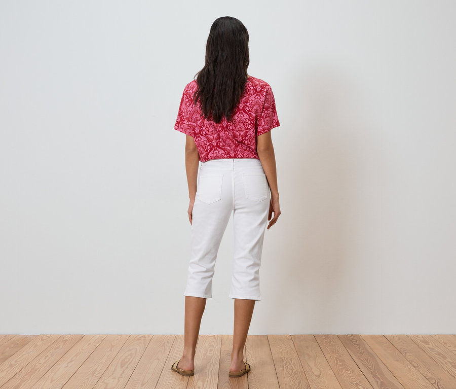 Eine Frau mit dunklen Haaren steht mit dem Rücken zur Kamera, trägt ein rot gemustertes Oberteil, weiße Dreiviertelhosen und goldene Sandalen.