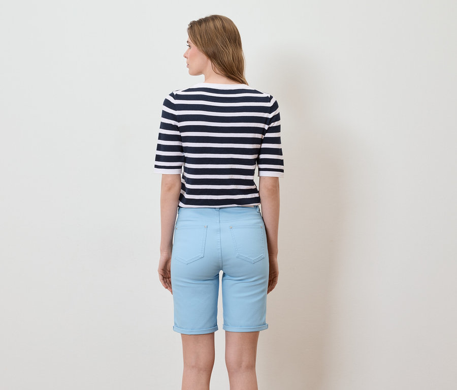 Rückansicht einer Frau mit langen, blonden Haaren, die ein gestreiftes marineblau-weißes Kurzarmshirt und hellblaue Shorts trägt.