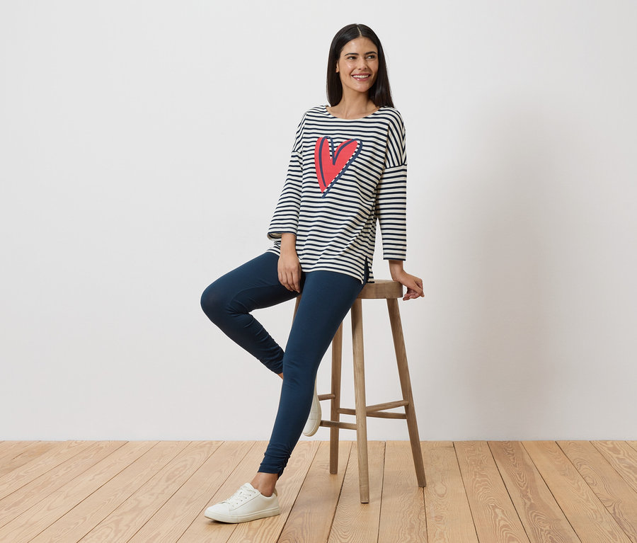 Frau sitzt auf einem Hocker, trägt ein gestreiftes Langarmshirt mit Herz, Leggings und weiße Sneaker.