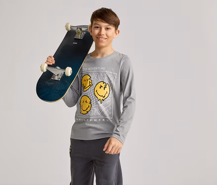 Lächelnder Junge hält ein Skateboard über der Schulter, trägt ein graues Langarmshirt mit gelben Smiley-Motiven und graue Hosen.