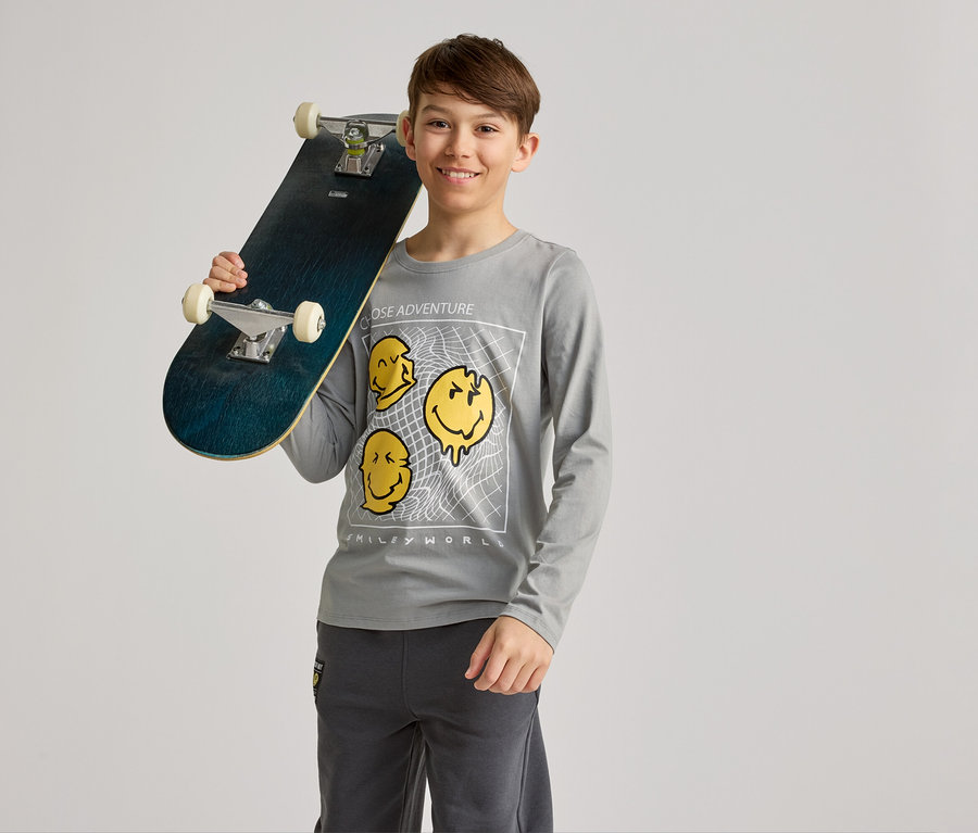 Lächelnder Junge hält ein Skateboard über der Schulter, trägt ein graues Langarmshirt mit gelben Smiley-Motiven und graue Hosen.