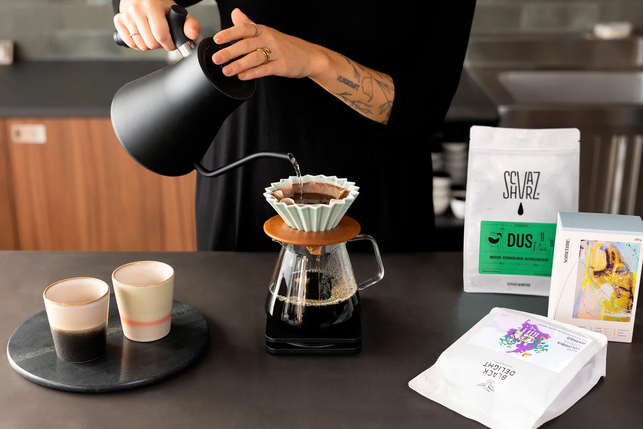 Frau gießt Wasser aus einem schwarzen Schwanenhalskessel in einen Kaffeefilter. Zwei Tassen und Kaffeepackungen stehen auf dem Tisch.