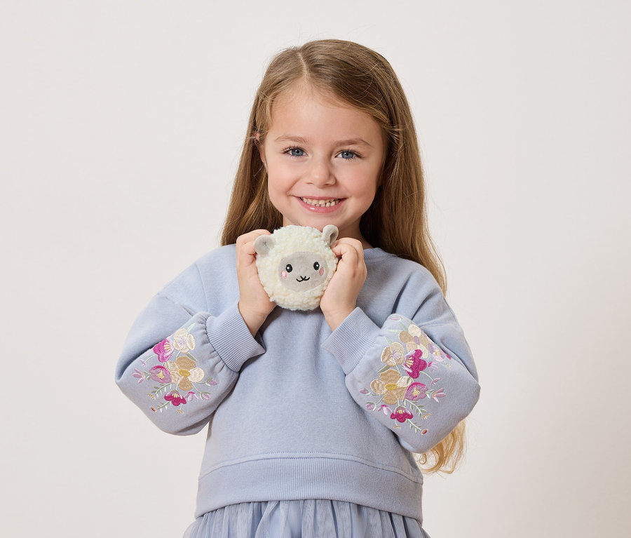 Ein junges Mädchen lächelt und hält ein Stofftier-Schaf vor sich. Sie trägt einen blauen Pullover mit Blumenstickereien an den Ärmeln.