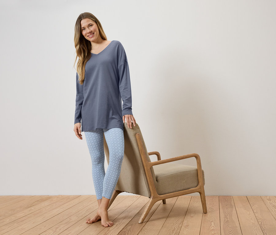 Frau mit langem, blauem Oberteil und hellblauen Leggings mit Muster, die sich an einen Stuhl mit beigem Sitz lehnt.