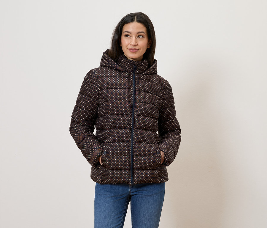 Frau mit brauner, gemusterter Daunenjacke mit Kapuze und blauen Jeans, die mit den Händen in den Taschen vor einer weißen Wand posiert.
