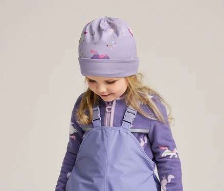 Mädchen mit blonden Haaren trägt eine lila Mütze mit Einhornmuster, eine Fleecejacke mit Reißverschluss und eine lila Latzhose.