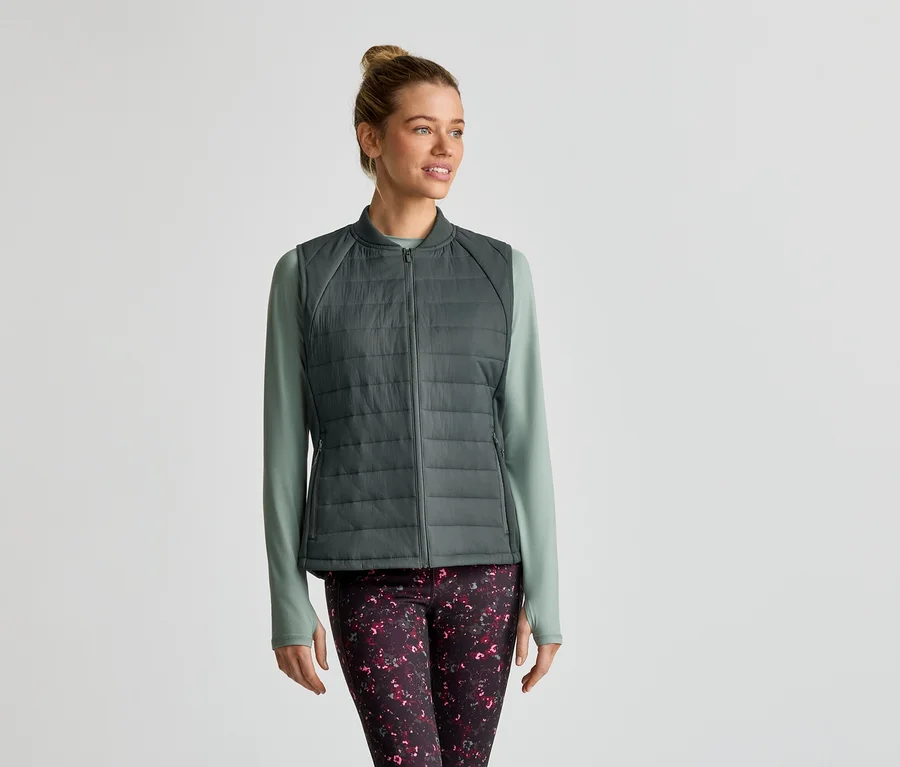 Frau mit Dutt trägt eine graue Steppweste über einem hellgrünen Langarmshirt und gemusterte Leggings.