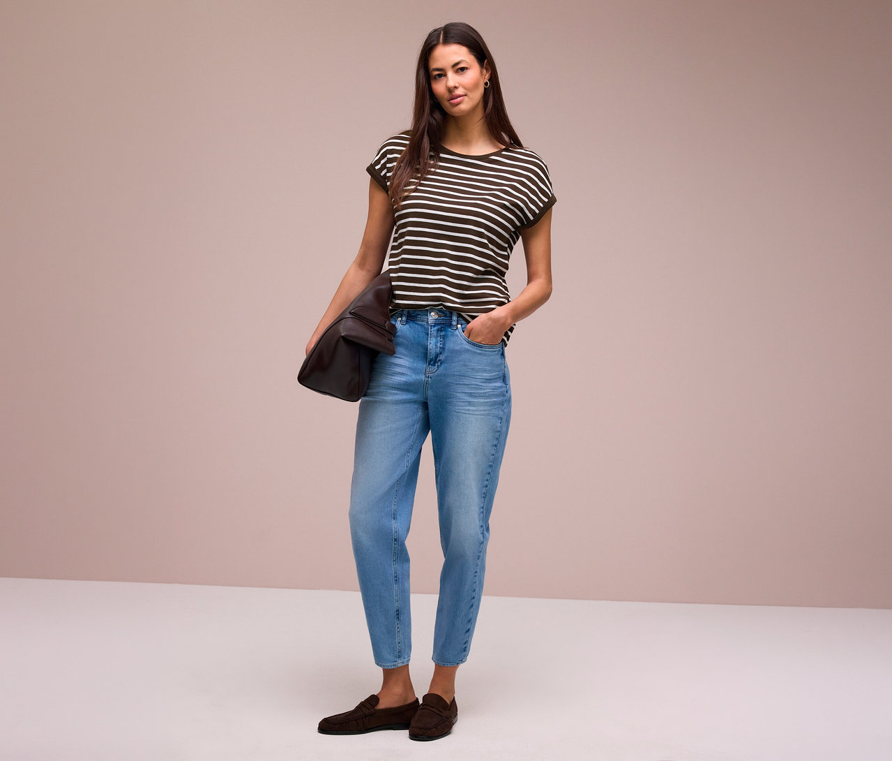 Eine Frau mit langen dunklen Haaren steht und blickt nach vorne. Sie trägt ein gestreiftes Street One T-Shirt, hellblaue Jeans und dunkelbraune Loafer und hält eine braune Tasche unter dem linken Arm.