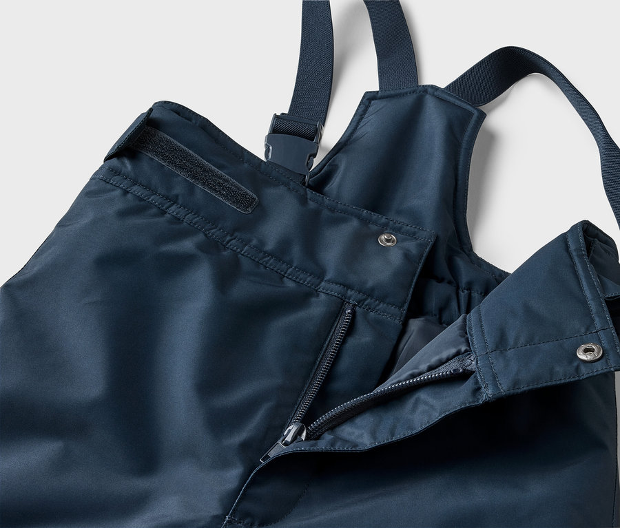 Nahaufnahme einer marineblauen Regenhose mit Trägern, teilweise geöffnetem Reißverschluss und silbernem Druckknopf.