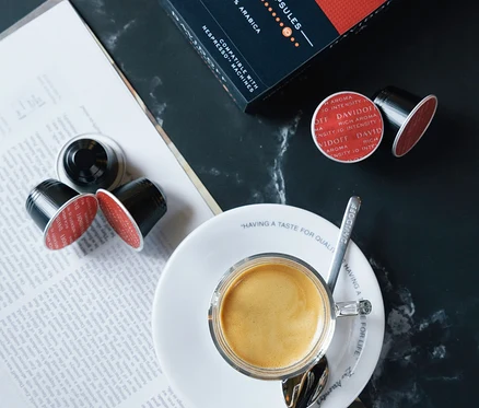 Espresso in Tasse auf Untertasse mit Löffel neben Packung DAVIDOFF CAFÉ Rich Aroma und Kaffeekapseln.