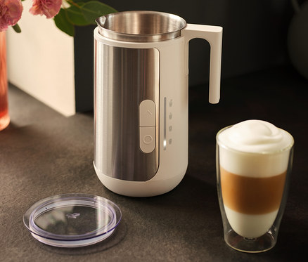Ein Milchaufschäumer aus Edelstahl steht neben einem Glas Latte Macchiato und einem Deckel auf einer dunklen Arbeitsfläche.