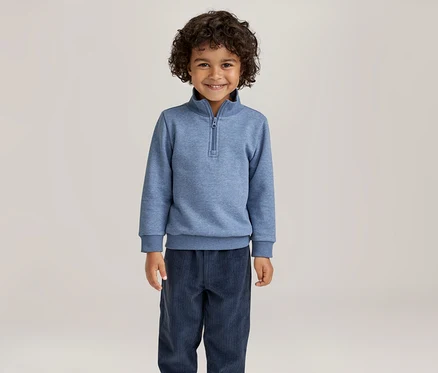 Lächelnder Junge mit lockigem Haar, der ein blaues Sweatshirt mit Reißverschlusskragen und eine dunkelblaue Hose trägt.