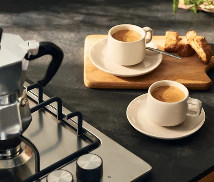 Silberne Mokkakanne auf einem Gasherd neben zwei hellen Kaffeetassen mit Untertassen und Biscotti auf einem Holzbrett.
