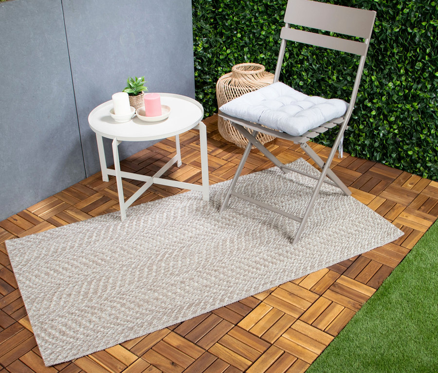 Ein hellbeiger Teppich »Twine«, natural flow, liegt auf einem Holzboden neben einem weißen Beistelltisch mit Kerzen und einem grauen Klappstuhl mit blauem Kissen.