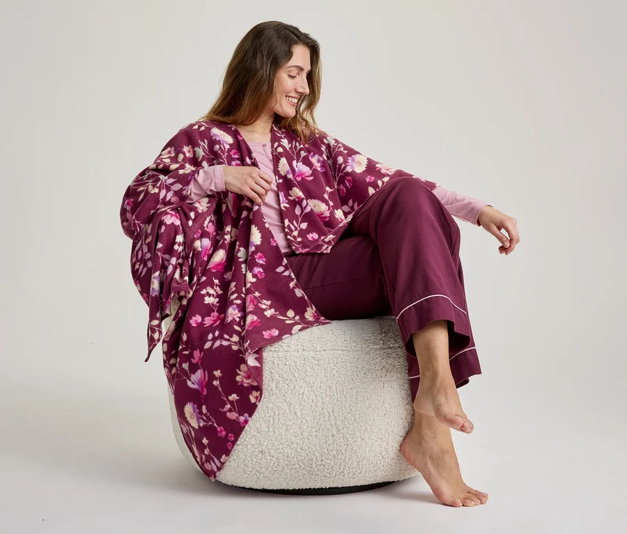 Eine lächelnde Frau mit langen braunen Haaren sitzt barfuß auf einem flauschigen weißen Pouf und trägt ein hellrosa Langarmshirt, eine bordeauxrote weite Hose mit rosa Zierleiste und einen bordeauxroten Kimono-Bademantel mit Blumenmuster.
