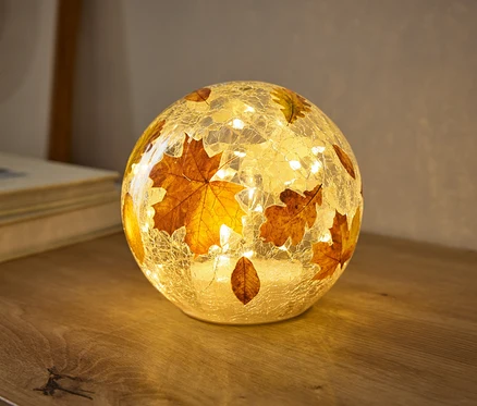 Eine kugelförmige Lampe mit rissiger Glasstruktur, verziert mit Herbstblättern, leuchtet warm auf einer Holzoberfläche. Kleine Lichter im Inneren beleuchten die Blätter.