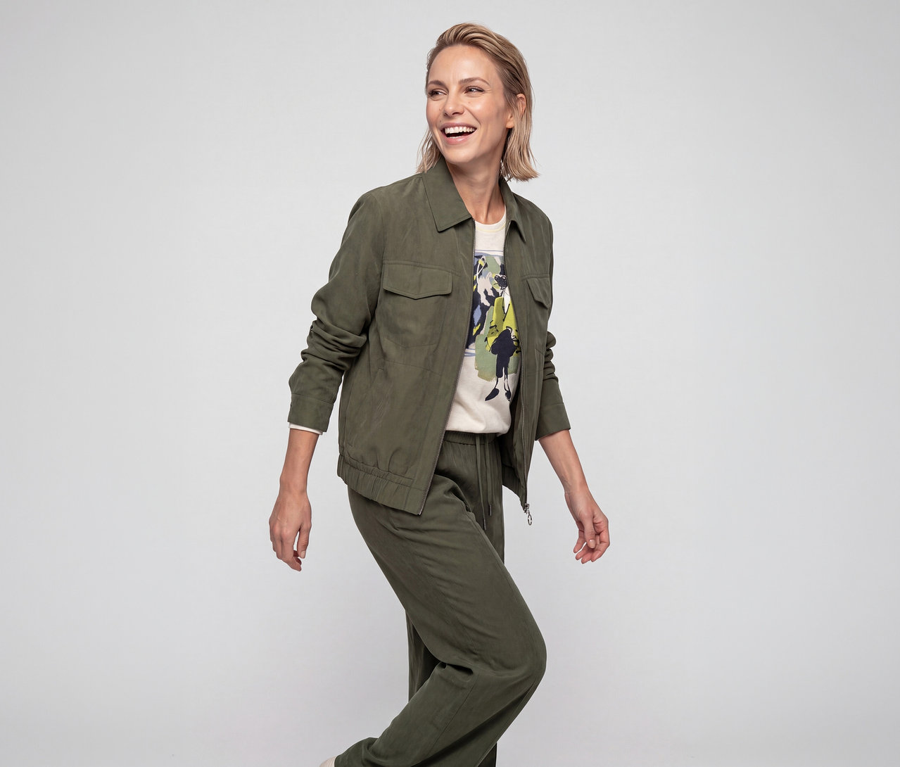 Lächelnde Frau mit kurzen blonden Haaren trägt eine grüne Jacke mit Brusttaschen, ein helles Grafik-T-Shirt und passende grüne Hose, blickt nach rechts.